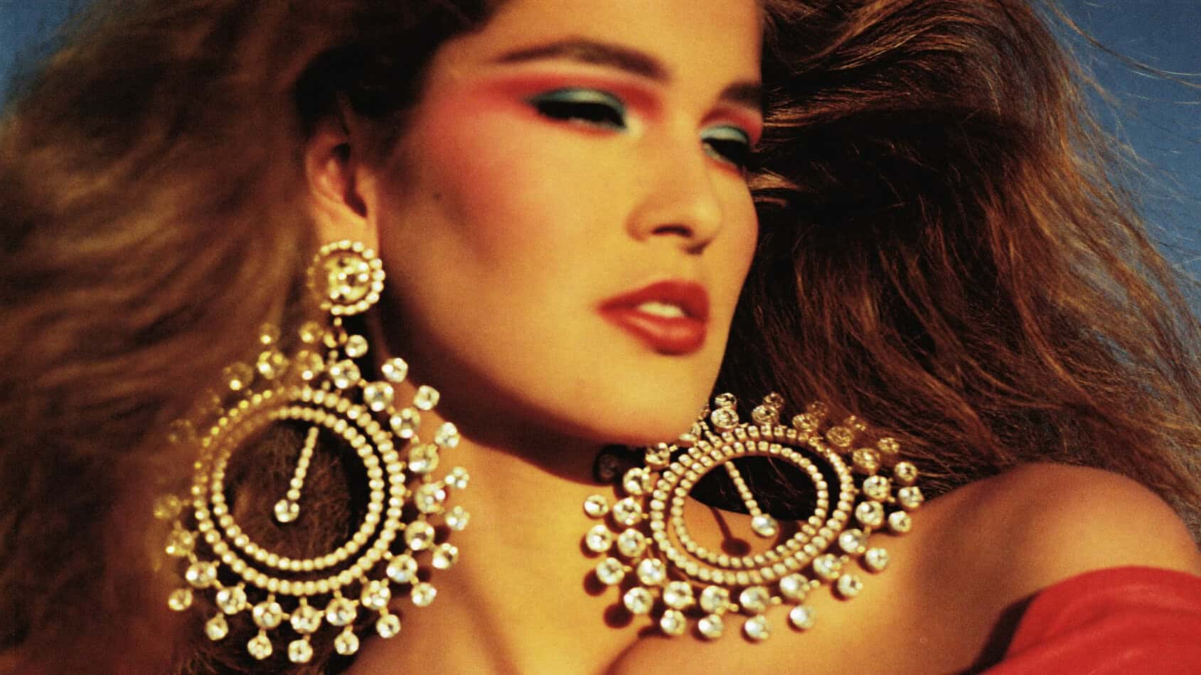 Close-up of glamorous 1980s woman wearing enormous gold chandelier earrings with rhinestones, bold makeup and voluminous hair