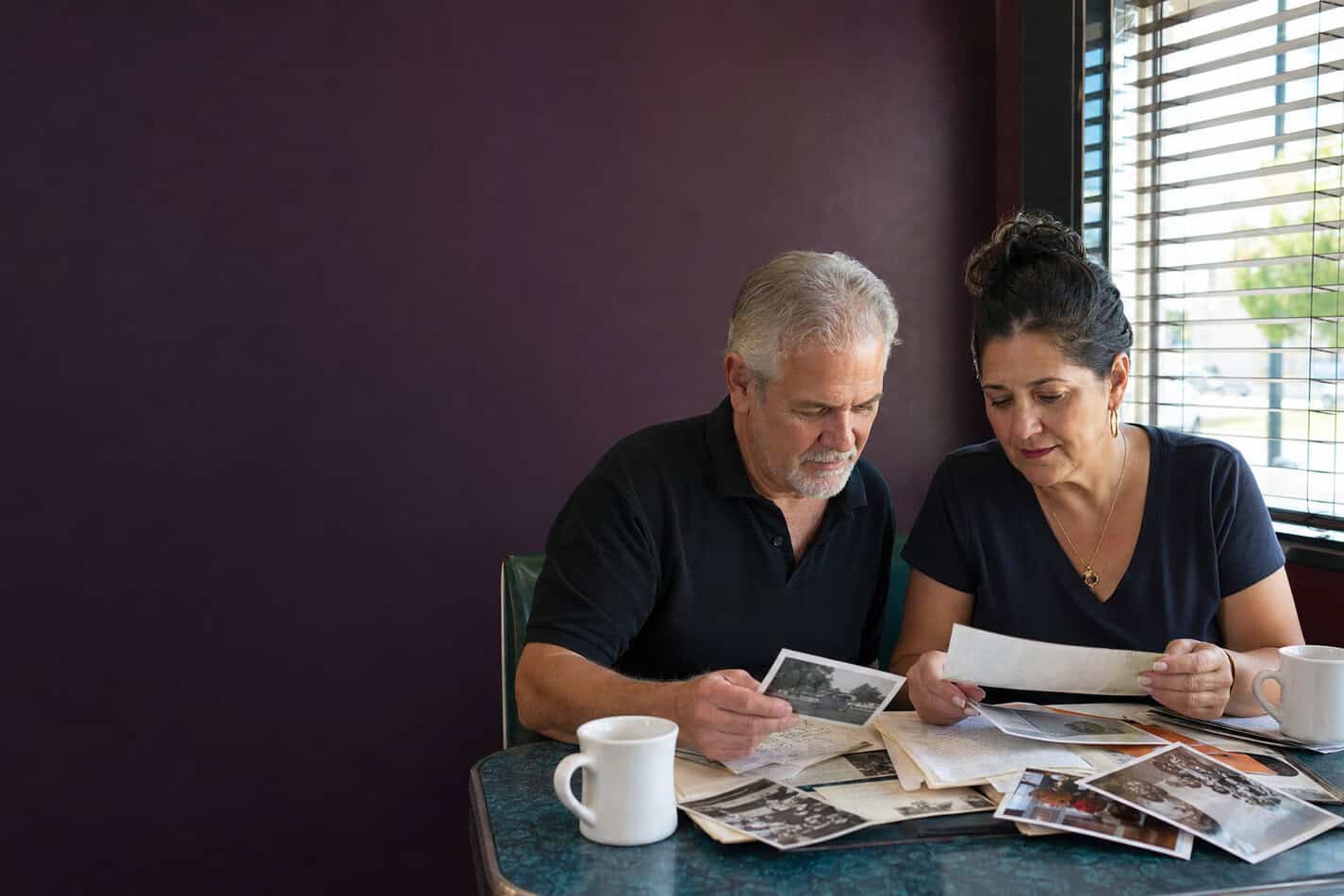 A business owner and a longtime customer sit together at a table, looking through old photographs. Natural light. Warm and unhurried.