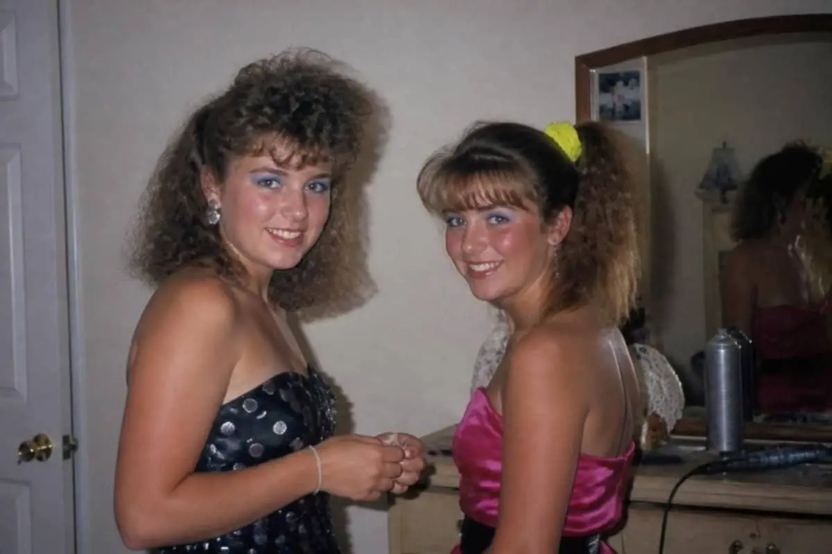 Two girls in the 1980s, one with a big curly perm and one with teased bangs and a side ponytail with a neon scrunchie