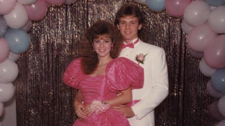 Couple in a hot pink puff sleeve 80s prom dress and white tux with matching pink cummerbund posed in front of a tinsel backdrop and balloons