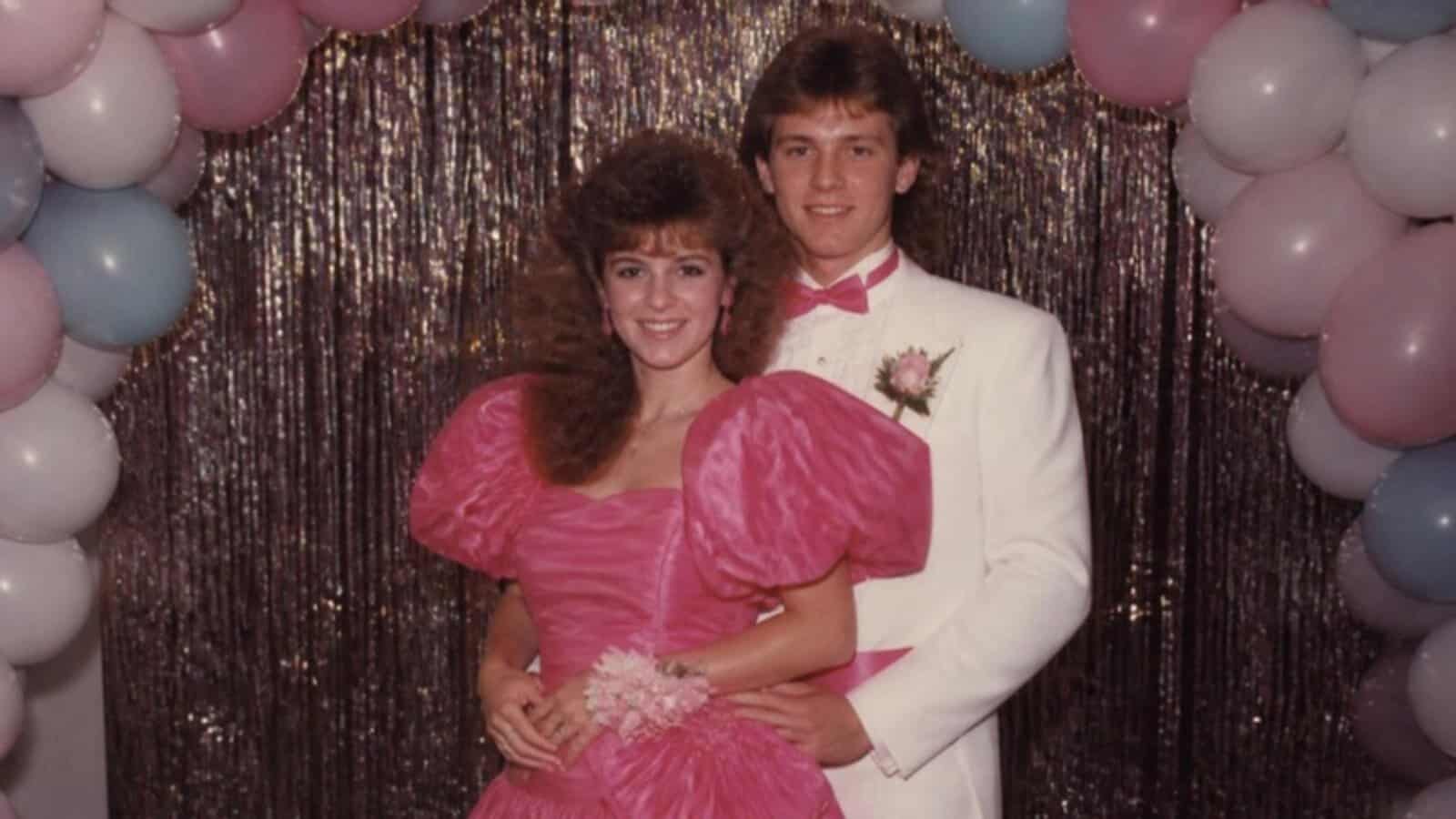 Couple in a hot pink puff sleeve 80s prom dress and white tux with matching pink cummerbund posed in front of a tinsel backdrop and balloons