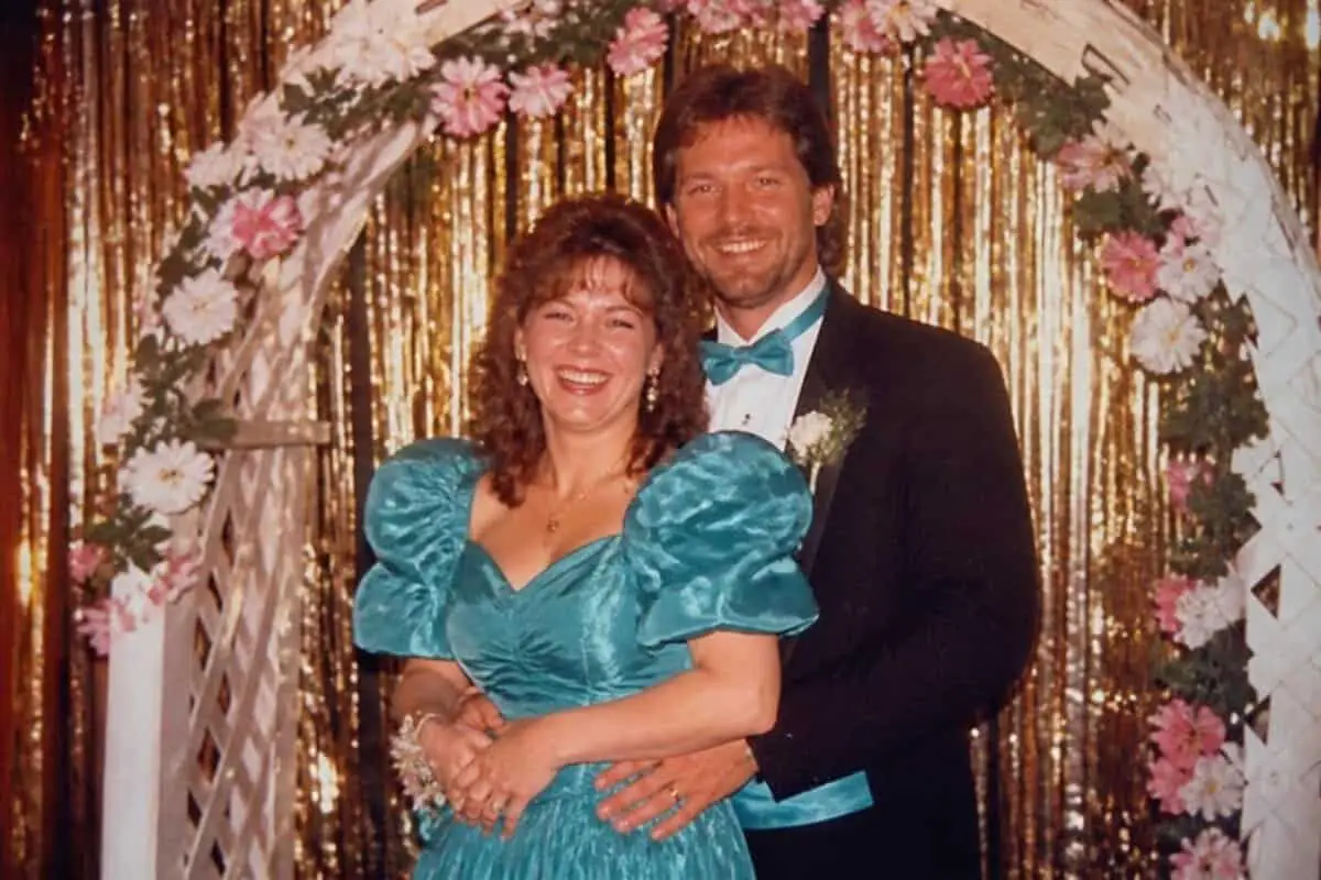 Couple recreating the classic 80s prom photo pose in a teal taffeta dress and tux under a flower lattice arch
