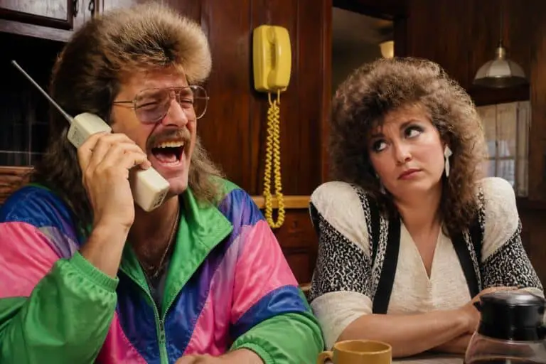 Funny 80s couple in a wood paneled kitchen, the husband with a mullet laughing into a giant brick cell phone while his wife rolls her eyes