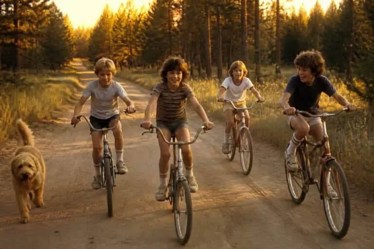 Four kids riding bikes down a dirt road at golden hour with a dog running alongside