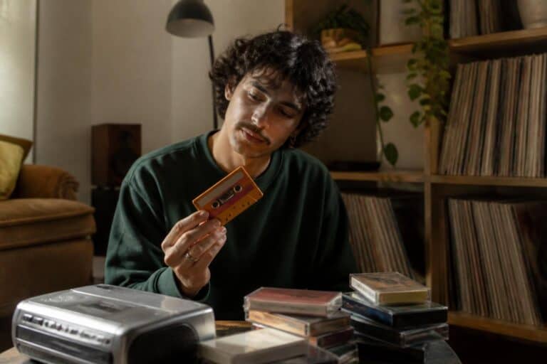 Young adult holding a colorful cassette tape next to a cassette player and a stack of tapes