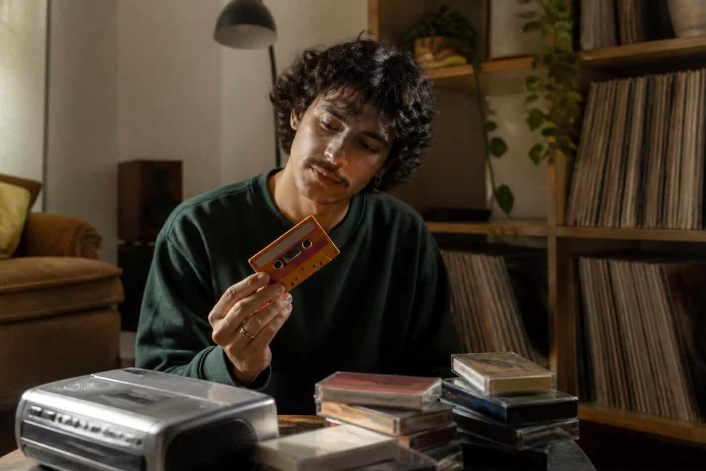 Young adult holding a colorful cassette tape next to a cassette player and a stack of tapes
