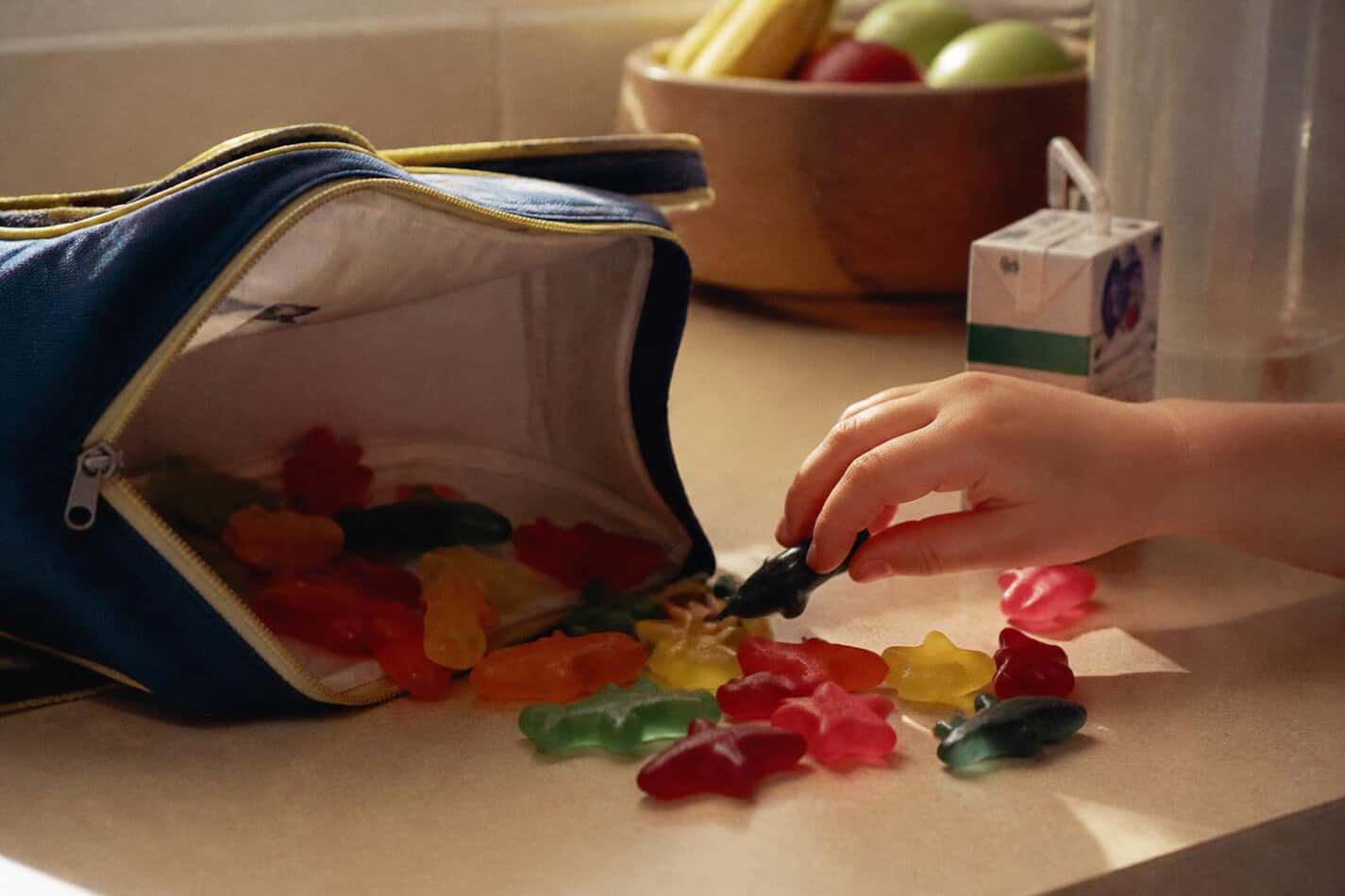 Lunchbox spilling colorful gummy fruit snacks next to a juice box, with a kid reaching in
