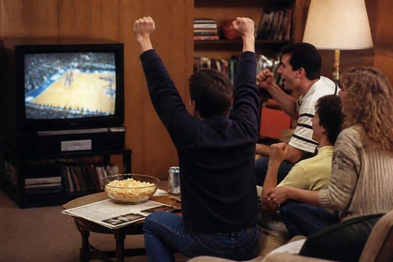 Friends cheering at a big box TV showing a basketball game in an 80s wood paneled living room