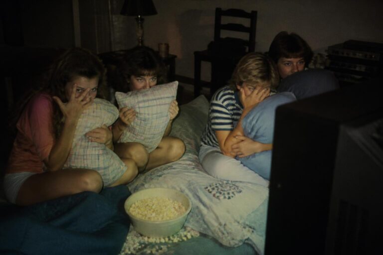 Teens at an 80s sleepover hiding behind pillows while watching a scary movie